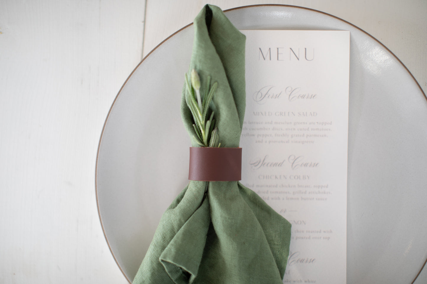 Minimal table setting featuring the Keyaiira Mahogany leather napkin ring on a white ceramic plate with a green linen napkin and printed menu  small batch crafted tableware for elegant, modern dining.