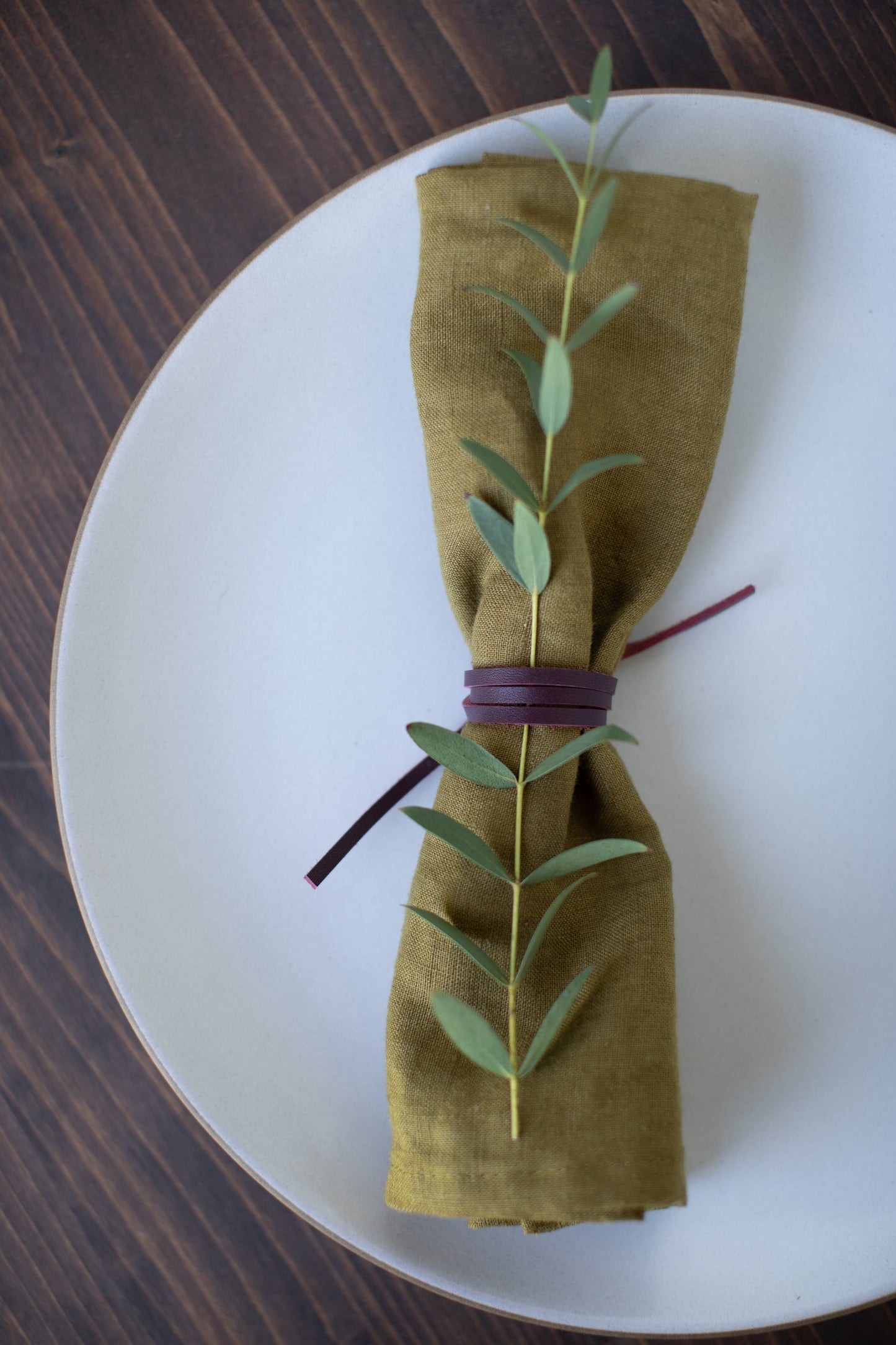 Wine-toned Leather Napkin Tie from Keyaiira. Soft, flexible leather cord wrapped around an earthy green napkin with eucalyptus sprig, adding a warm, modern touch to elegant dining decor.