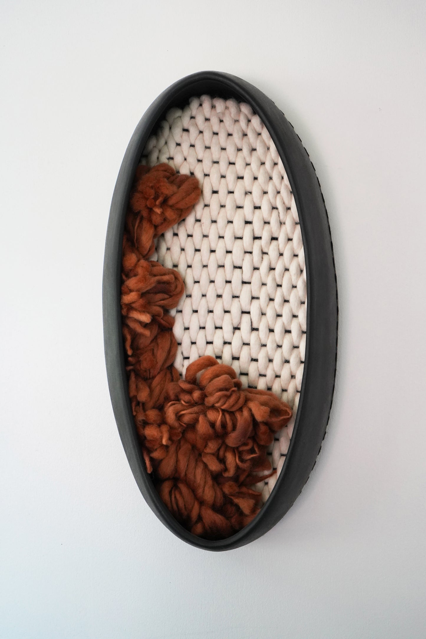 Sustainable wall decor made from ethically sourced materials. Matte black ceramic vessel filled with hand-dyed rust wool yarn and ivory hand-felted fibers. Sophisticated California design.