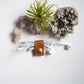 A white wired earbud holder with a brown leather strap and gold snap button, holding white earbuds, arranged on a white surface with crystals and a small air plant.