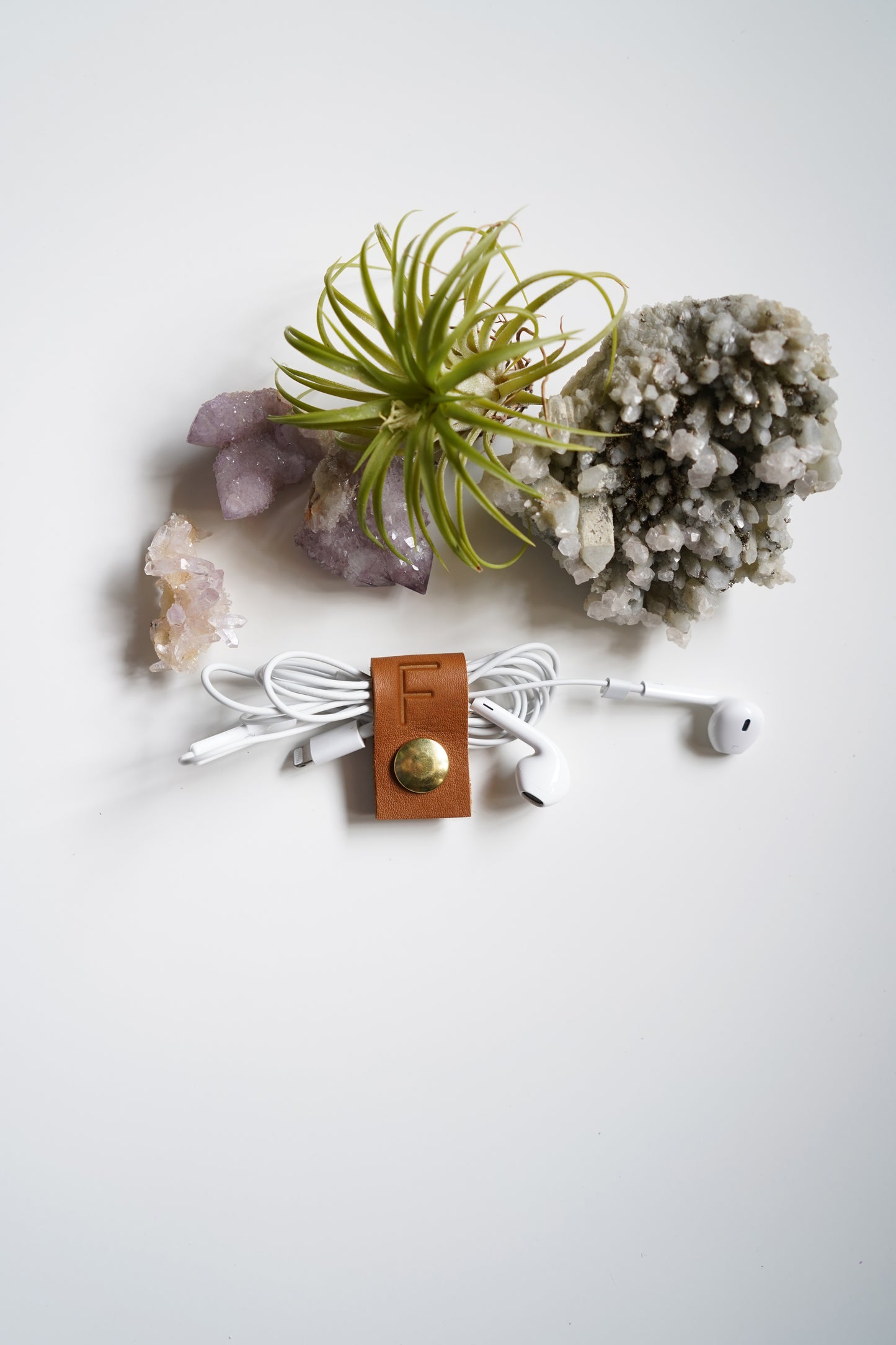 A white wired earbud holder with a brown leather strap and gold snap button, holding white earbuds, arranged on a white surface with crystals and a small air plant.