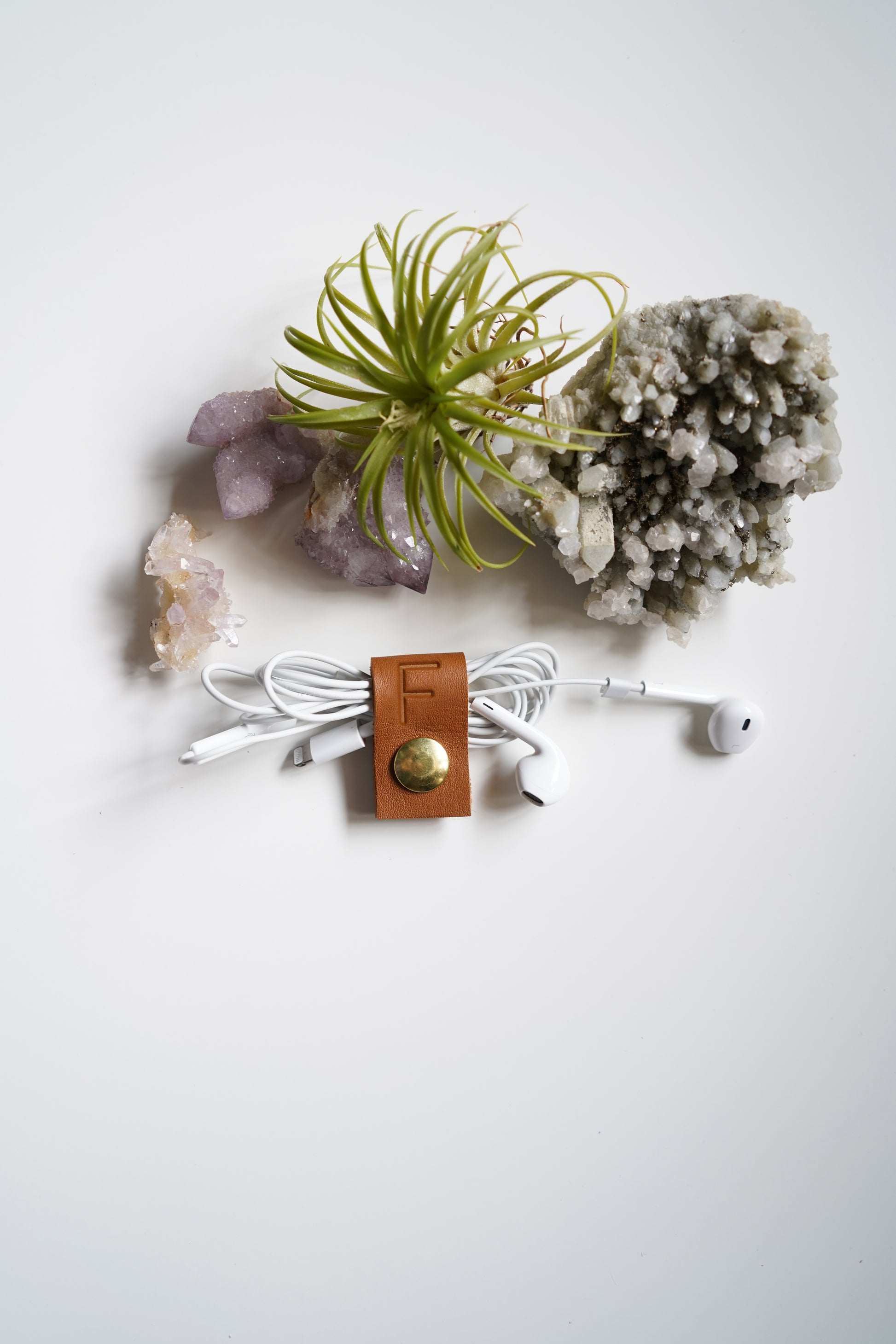 A white wired earbud holder with a brown leather strap and gold snap button, holding white earbuds, arranged on a white surface with crystals and a small air plant.