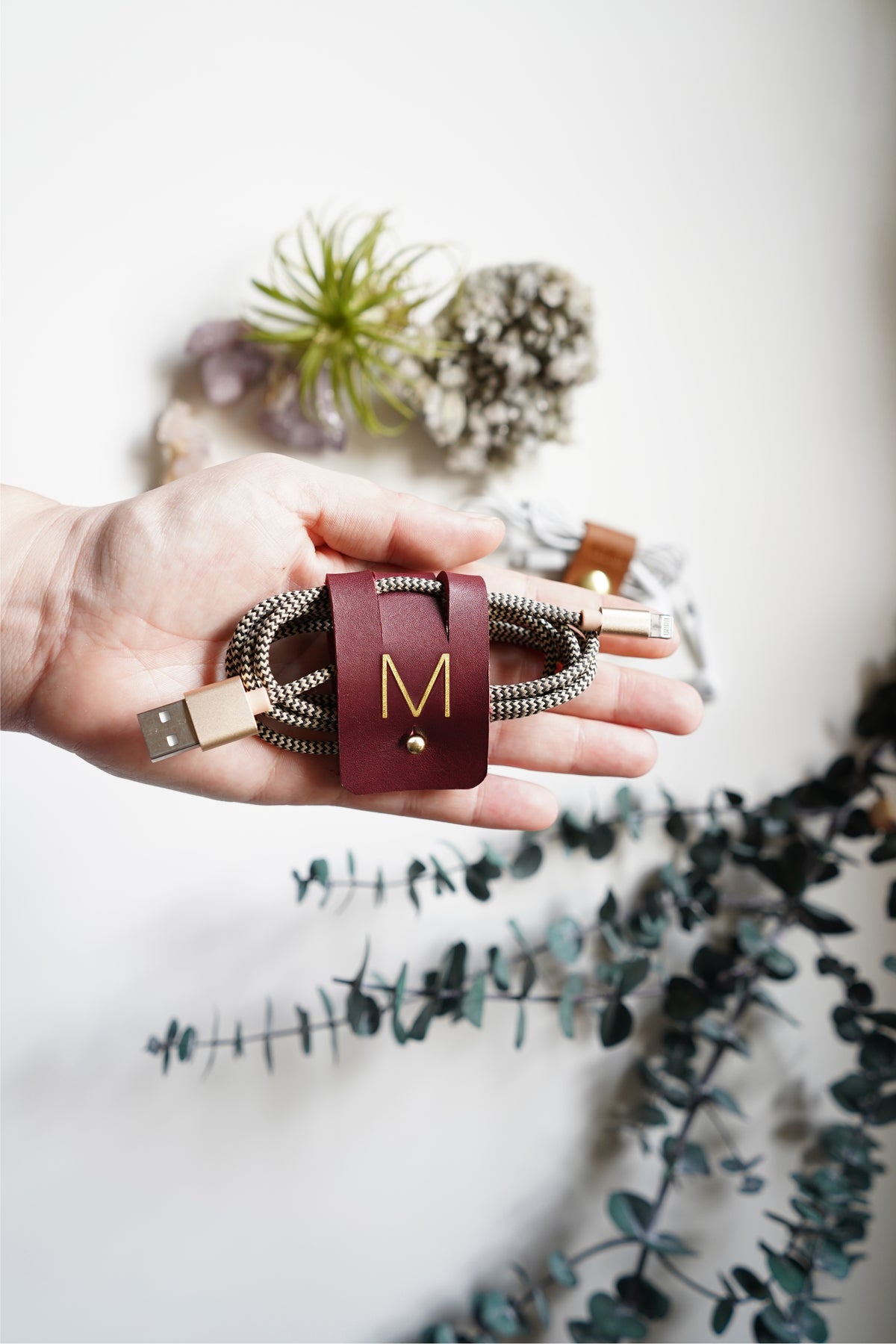 Wine leather cable keeper with personalized M initial securing a braided charging cord, carefully crafted by Keyaiira for USB cables, mobile electronics, travel tech and cable management
