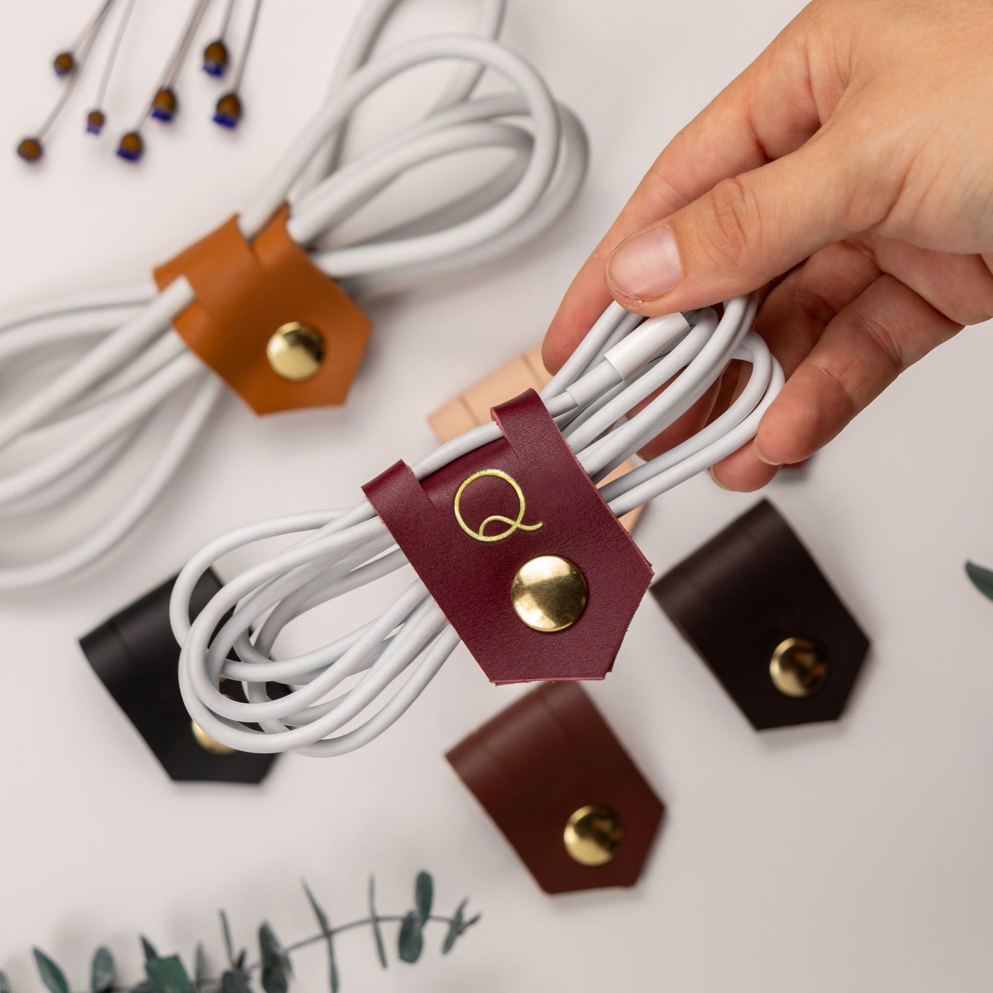 A hand holds a white cable secured by a burgundy leather cable organizer with a gold snap button and a stylized Q logo. Other cable organizers in brown, black, and tan are scattered on a white surface with decorative eucalyptus leaves.