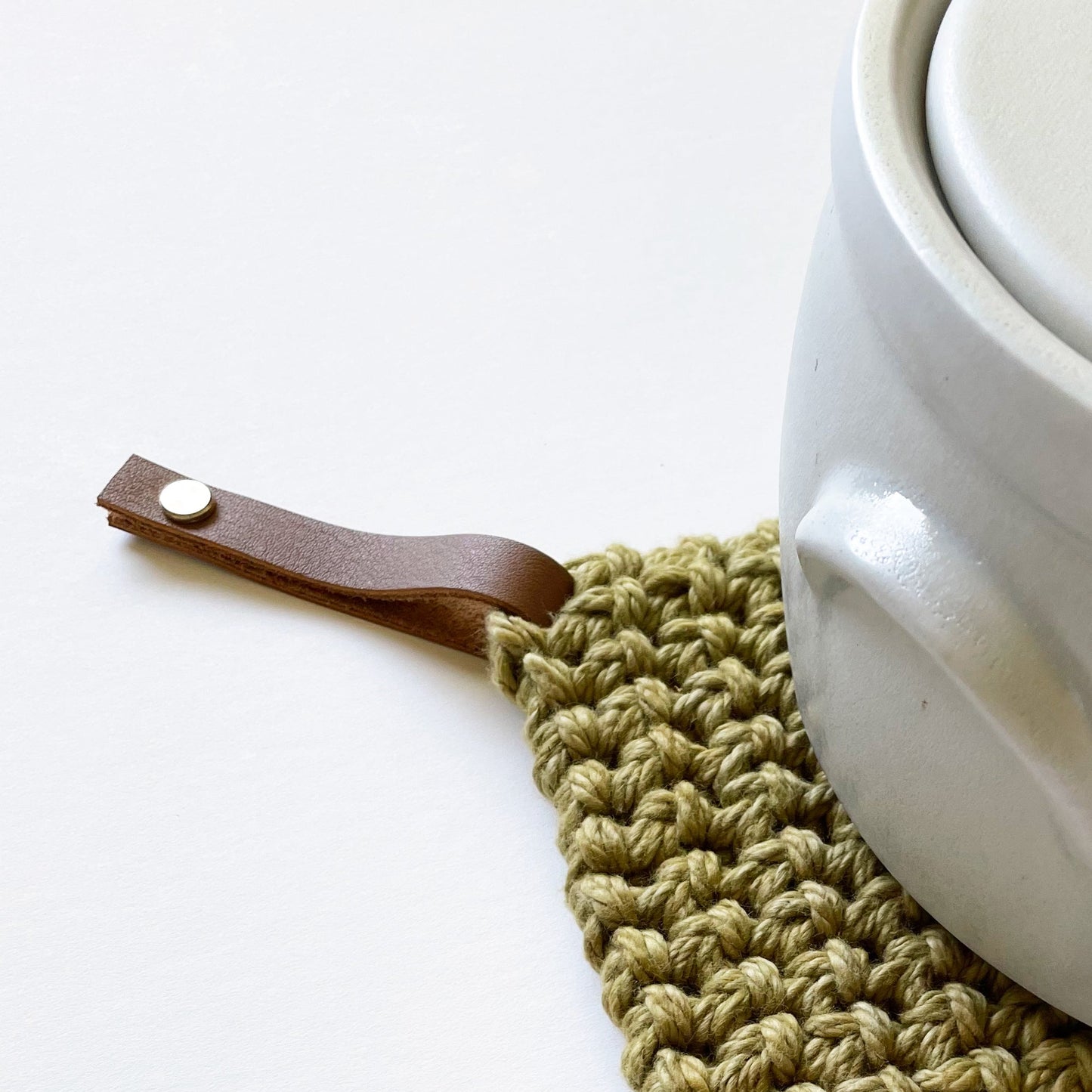 White ceramic pot with a brown leather handle on a green crocheted trivet.