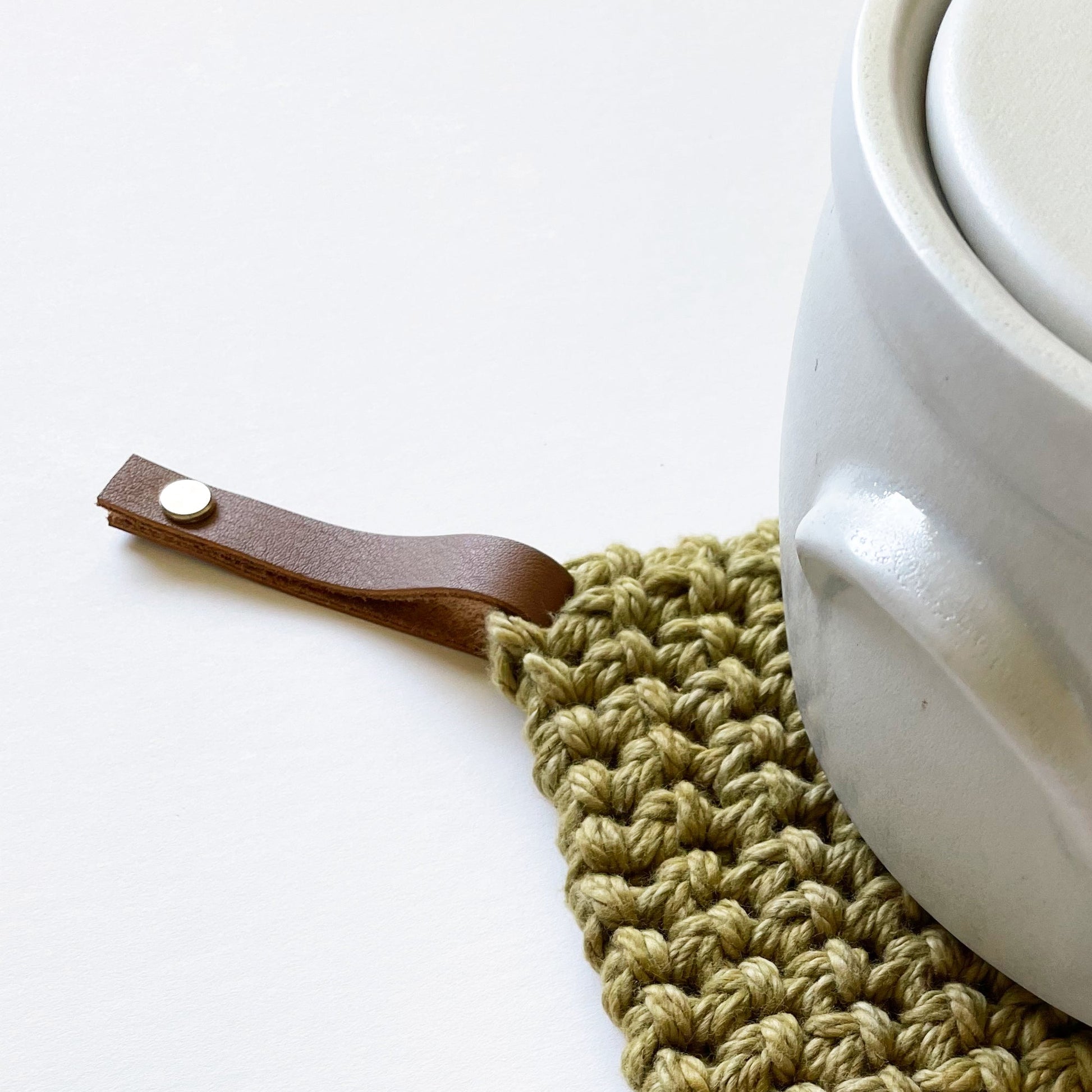 White ceramic pot with a brown leather handle on a green crocheted trivet.