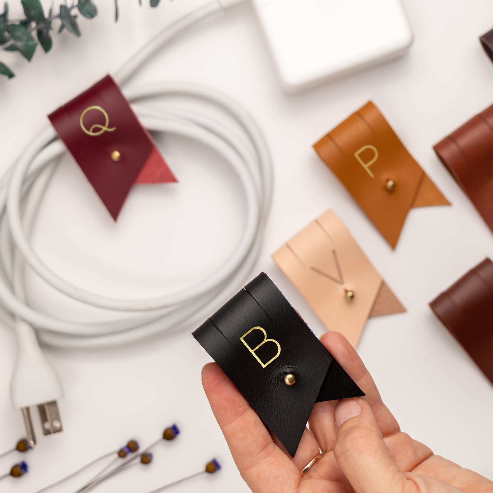Custom letter leather cable organizers in Wine, Black, Veg Tan, Nicotine and rich brown Keyaiira colors, designed for charger cords, earbuds, electronics and portable cable storage
