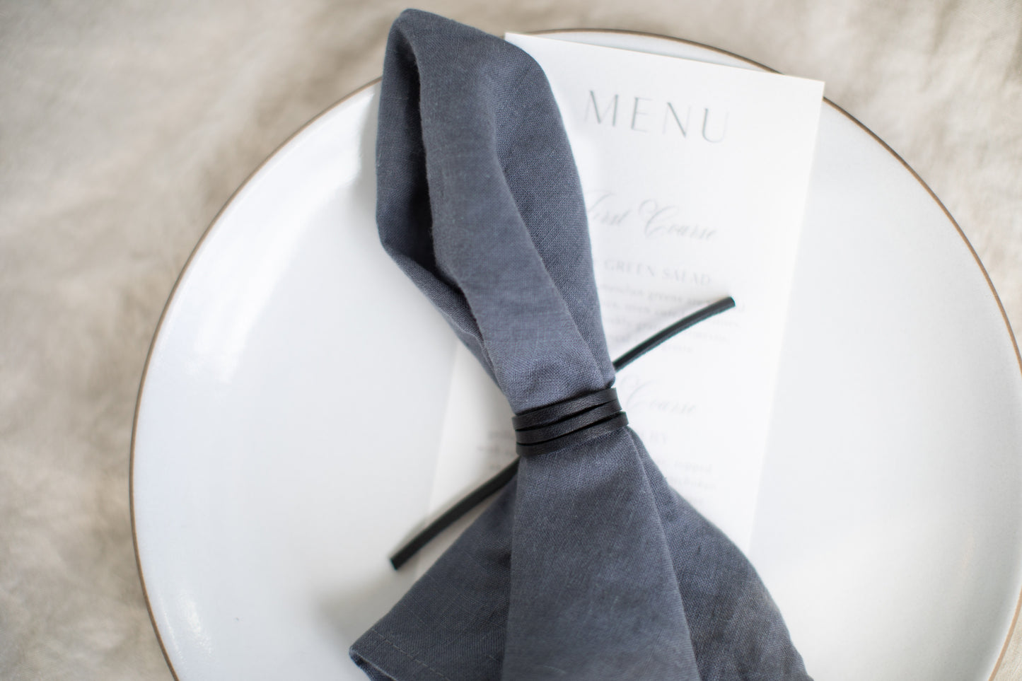 Leather Napkin Tie in black wrapped around a soft grey linen napkin with a printed menu card, minimalist, artisan-made table accessory crafted from sustainable leather for refined, modern dining decor by Keyaiira.