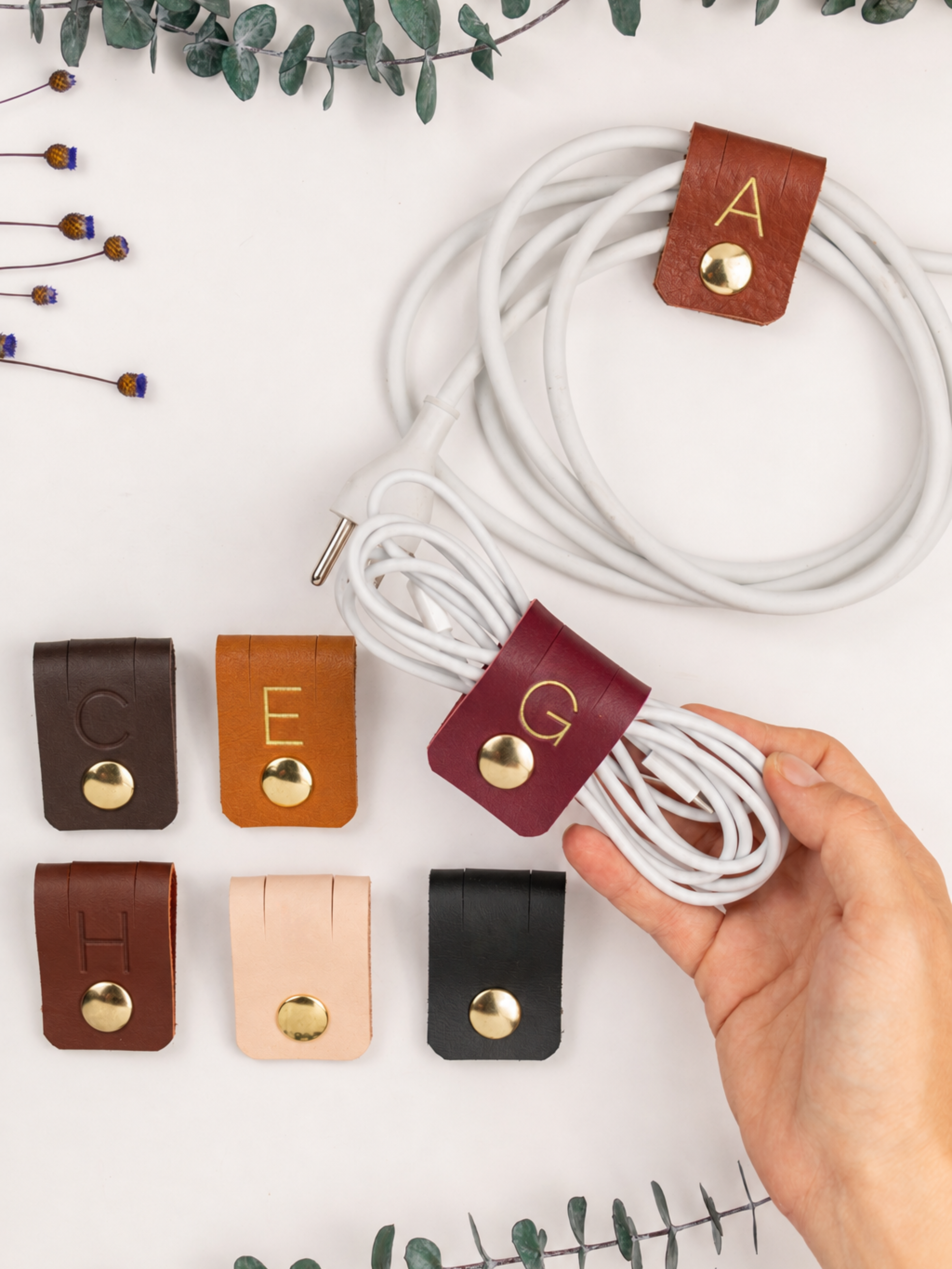 A hand holds a coiled white cable secured with a maroon leather cable tie featuring a gold snap and the letter G. Six other cable ties in brown, tan, blush, and black are arranged below, and the main cable tie with letter A is shown above on a white background with decorative greenery.