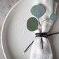 Close-up of a Keyaiira leather napkin tie wrapped around a soft linen napkin, styled on a white ceramic plate with eucalyptus greenery — handcrafted leather napkin holder for minimalist, Scandinavian-inspired table setting and modern dining decor.