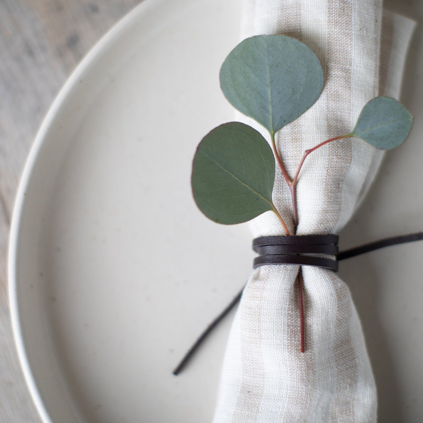 Close-up of a Keyaiira leather napkin tie wrapped around a soft linen napkin, styled on a white ceramic plate with eucalyptus greenery — handcrafted leather napkin holder for minimalist, Scandinavian-inspired table setting and modern dining decor.