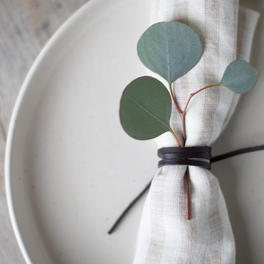 Close-up of a Keyaiira leather napkin tie wrapped around a soft linen napkin, styled on a white ceramic plate with eucalyptus greenery — handcrafted leather napkin holder for minimalist, Scandinavian-inspired table setting and modern dining decor.