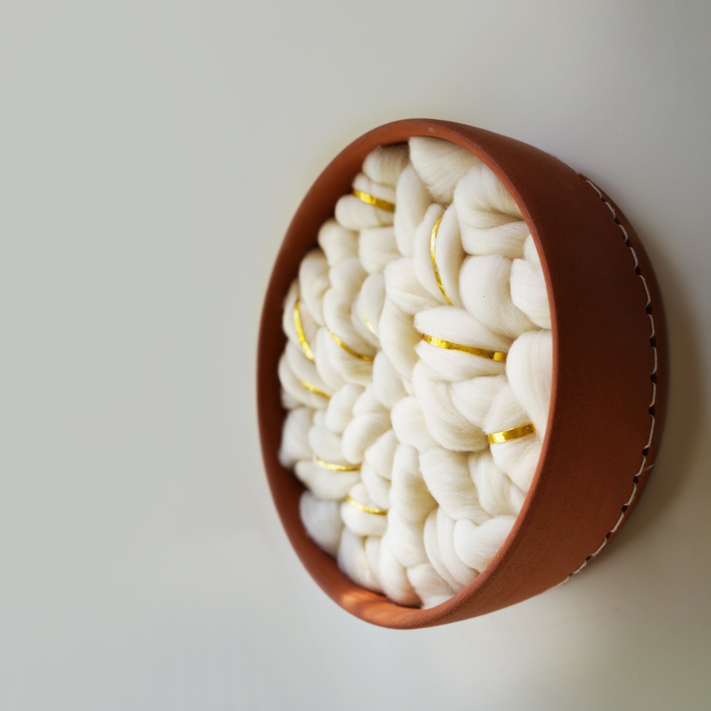 Round Handcrafted brown terracotta clay wall hanging, sized 12 inches. The interior features cloud-like white Rambouillet wool roving with delicate gold metal accents woven throughout. Modern organic fiber art by Keyaiira.