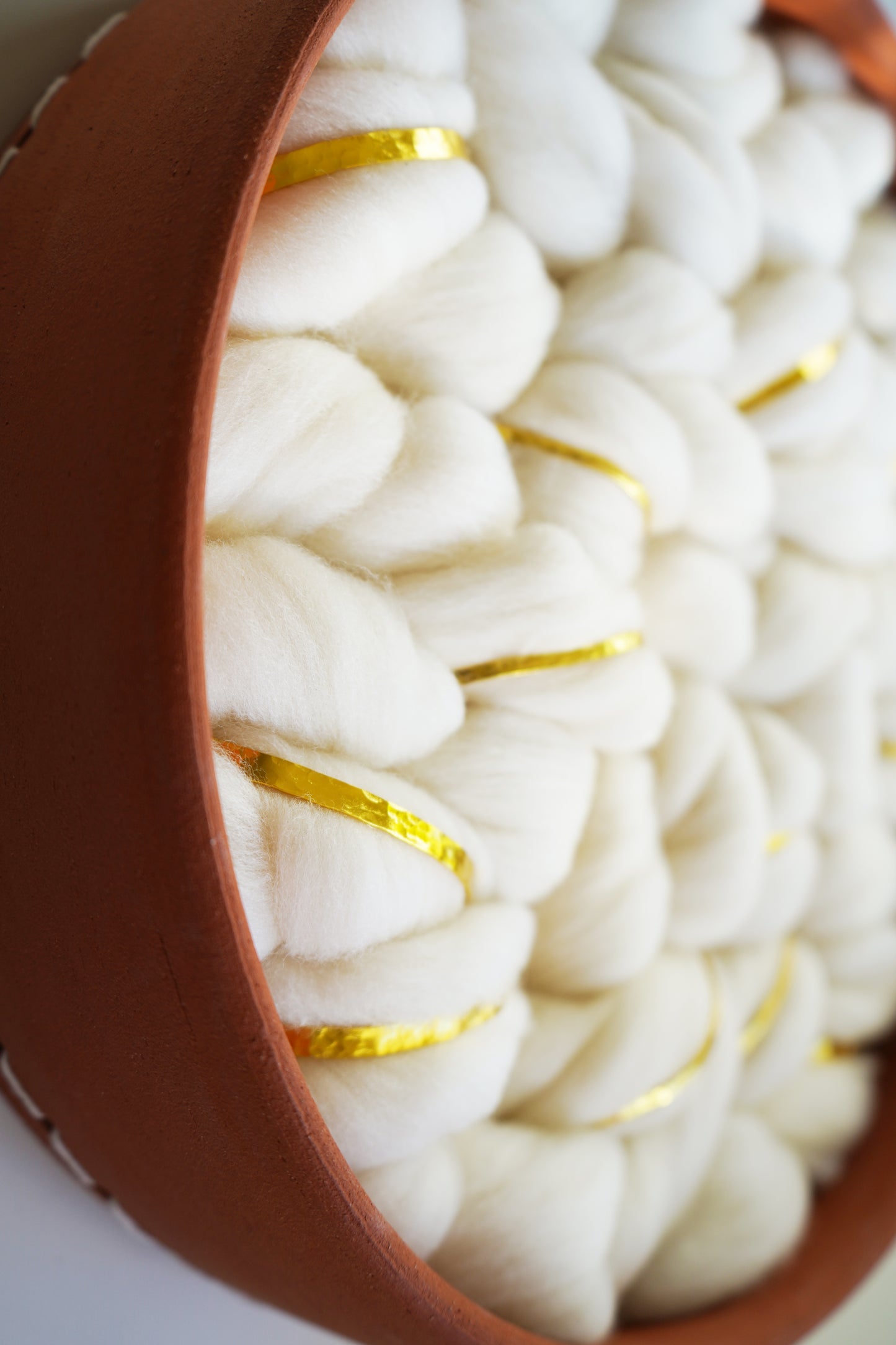 Macro detail of Fibershed Certified™ Rambouillet wool roving showing the soft, raw texture. subtle gold metal strands are woven into the white fibers, creating a shimmer against the matte terracotta clay rim.