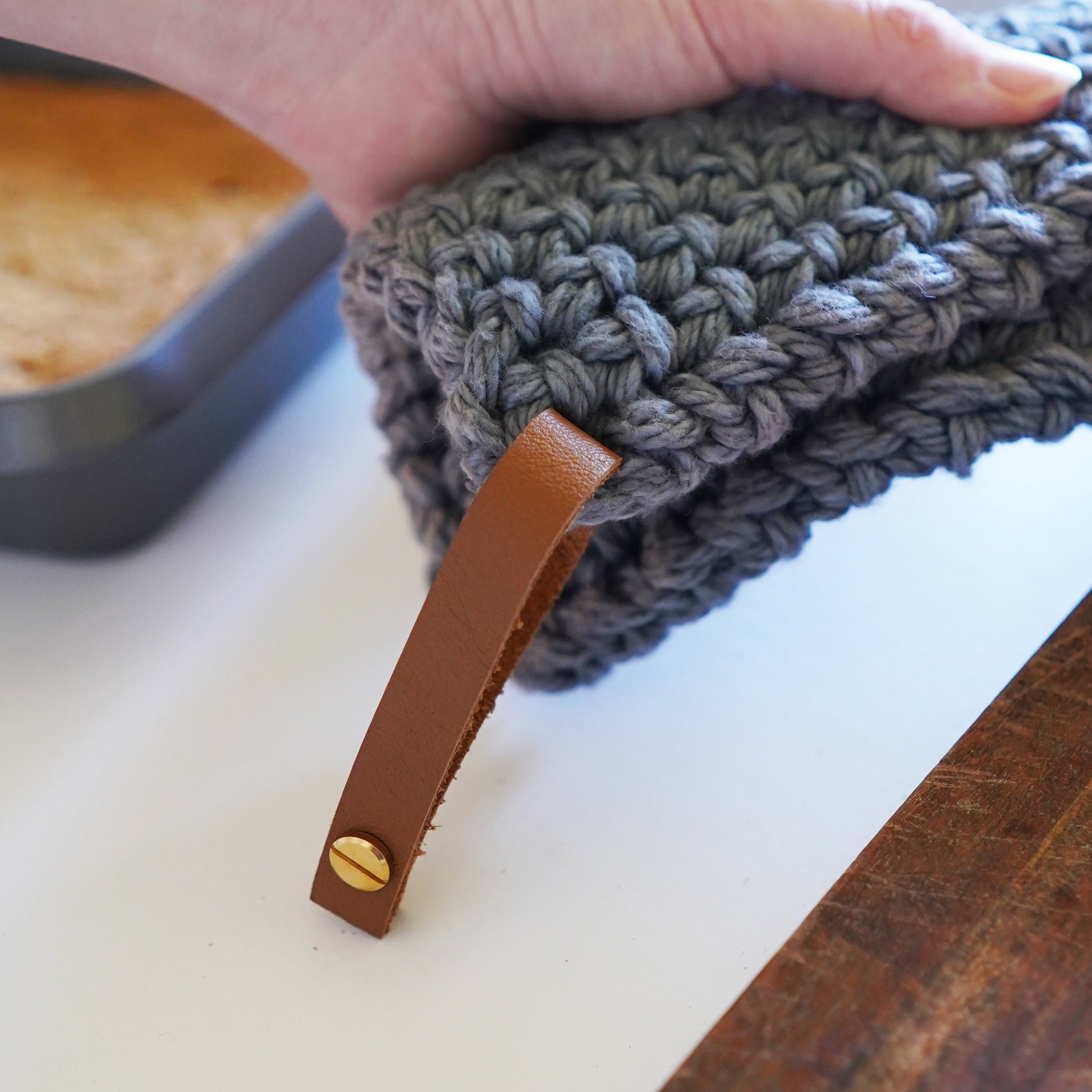 Person holding a blue-gray crocheted knit potholder with a detachable leather hanging loop over a wooden cutting board — handcrafted Keyaiira kitchen potholder with removable leather loop.