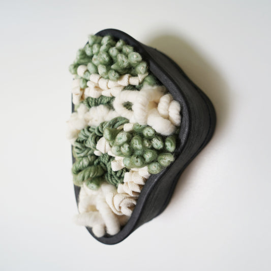 Full view of a handcrafted sculptural wall hanging featuring a brushed matte black clay frame. Soft, intricate fibers made from recycled plastic bottles and wool extend slightly beyond the ceramic edge, contrasting with the rigid frame.