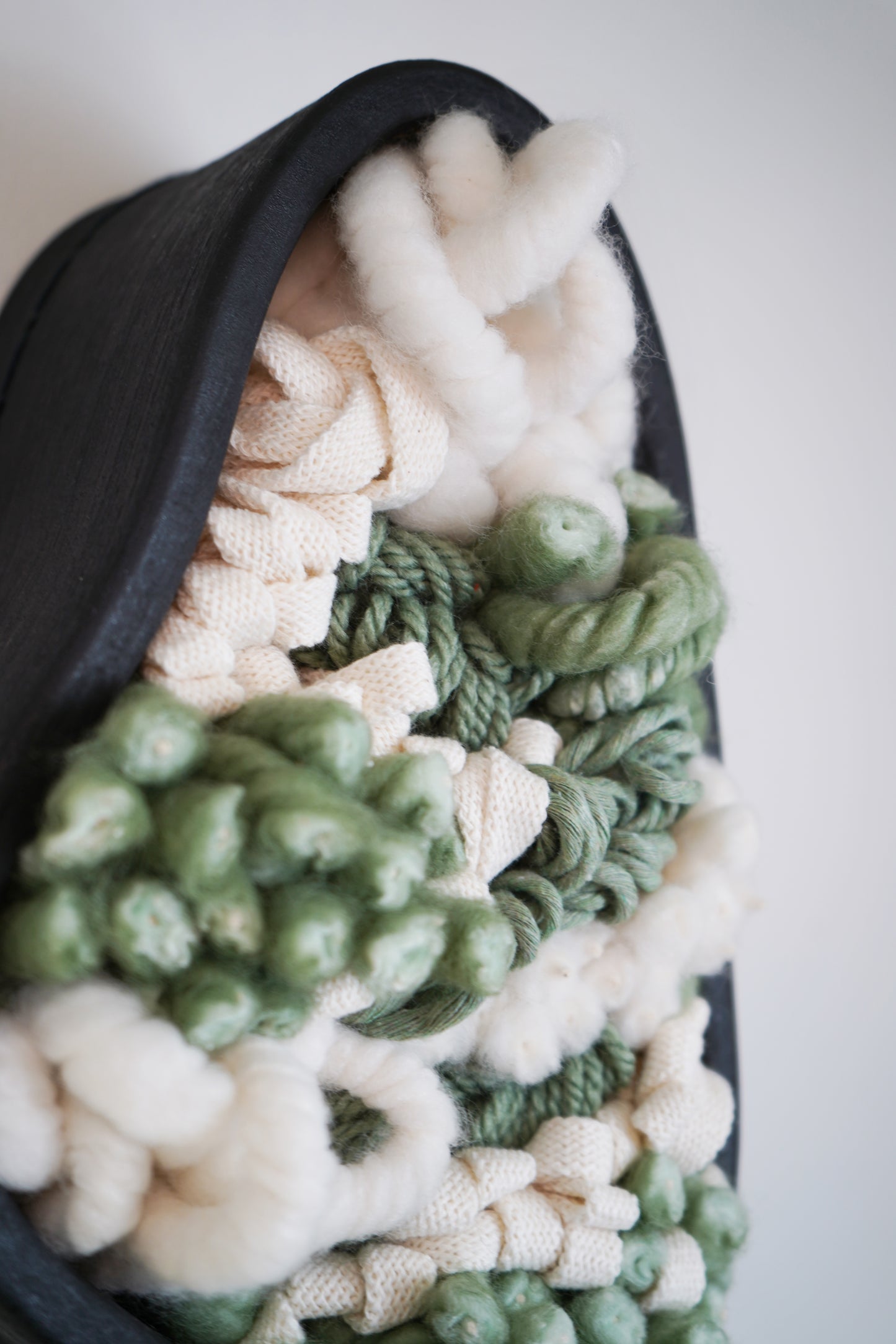 Macro detail shot showing the high contrast between the smooth, rigid brushed black clay and the soft, fuzzy texture of light-colored wools. Highlights the tactile quality of the sustainable materials used in this mixed-media wall sculpture.