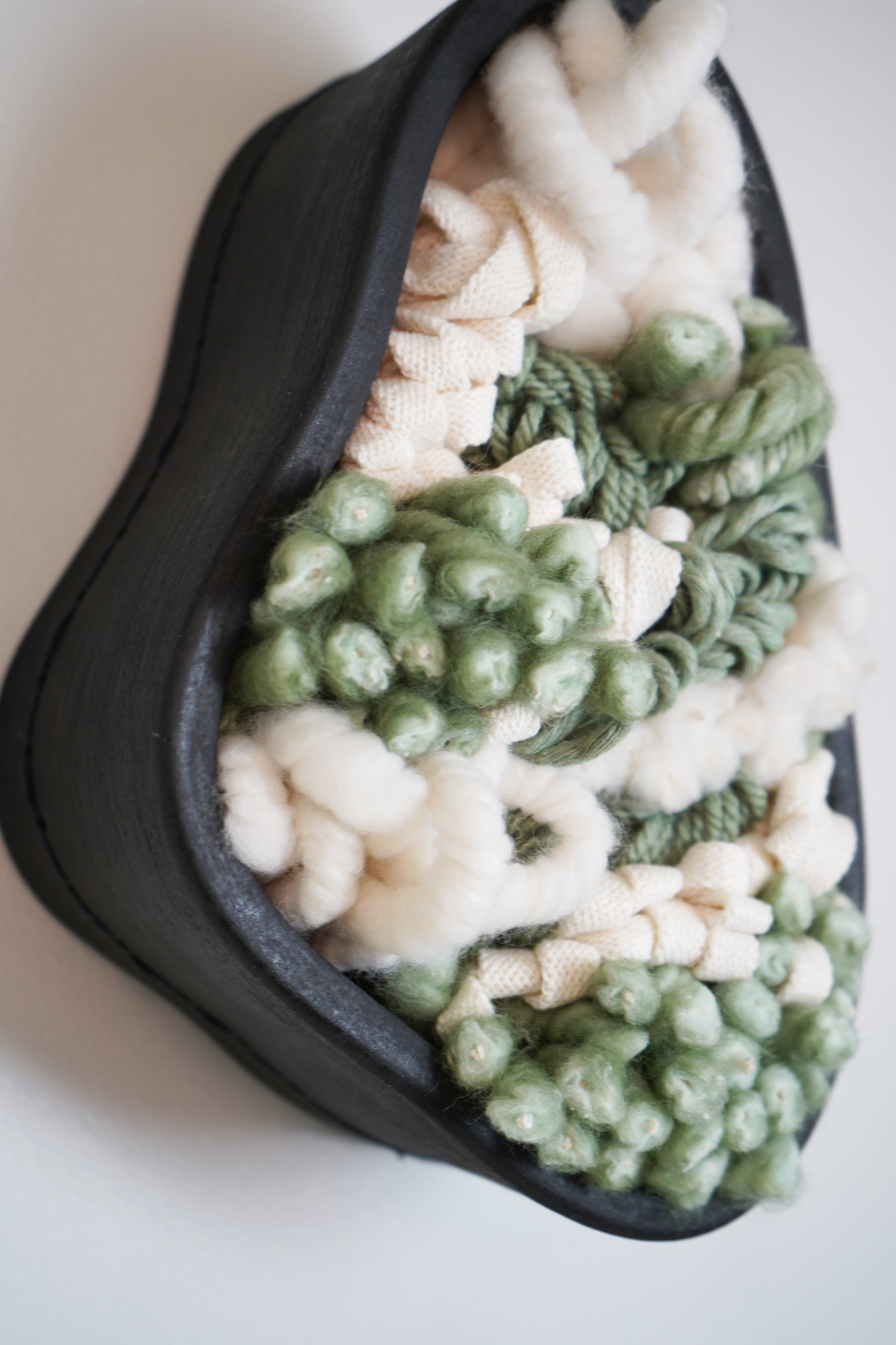Handcrafted wall sculpture featuring a brushed black terracotta clay frame filled with soft, textured fibers in sage green, aloe green, and cream tones. The interior is densely woven with sustainable materials including organic cotton, ethically sourced merino wool, braided Egyptian cotton, and yarns spun from recycled plastic bottles. This modern botanical wall art blends natural texture with sculptural depth, making it ideal for organic home decor, fiber art lovers, or those seeking unique artisan art