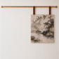 A rectangular wall art print with a sepia-toned landscape scene hangs from two brown leather straps on a gold horizontal rod against a plain off-white wall.