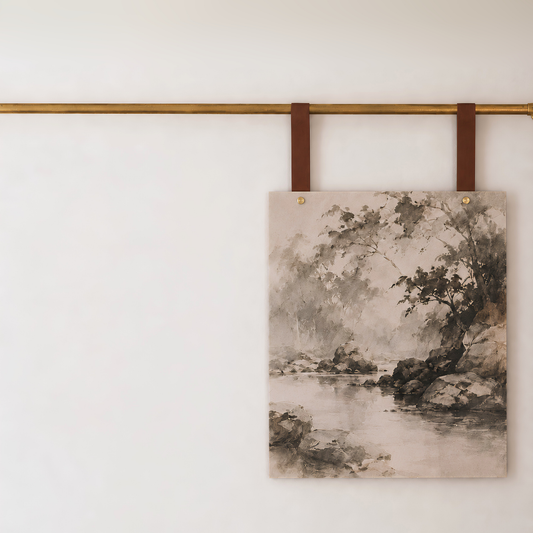 A rectangular canvas wall art print featuring a monochrome landscape painting of trees and water, hanging from two brown leather straps on a brass rod against a white wall by keyaiira.