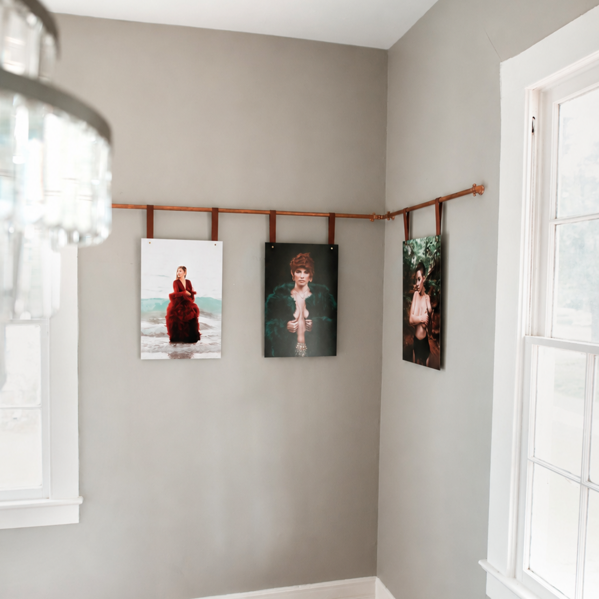 Three vertical art prints displayed on a wooden hanging rod strung across a gray wall in a room with hardwood floors and white-trimmed windows.