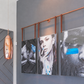 Three vertical framed art prints with mixed black-and-white portraits and bright blue graphic accents, hung on a gray wall under a copper-colored metal rod. A copper pendant light hangs to the left.