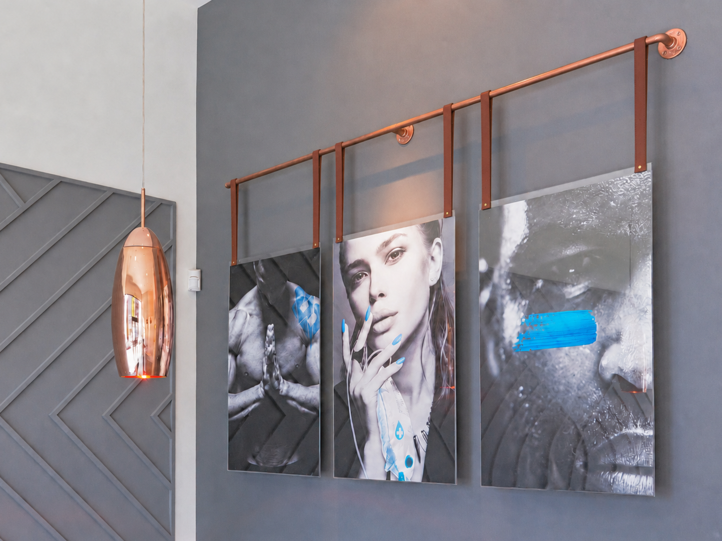 Three vertical framed art prints with mixed black-and-white portraits and bright blue graphic accents, hung on a gray wall under a copper-colored metal rod. A copper pendant light hangs to the left.