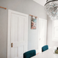 A styled interior room with gray walls, white paneled doors, a crystal chandelier, a dining table, teal chairs, and framed wall art hanging from copper rods.