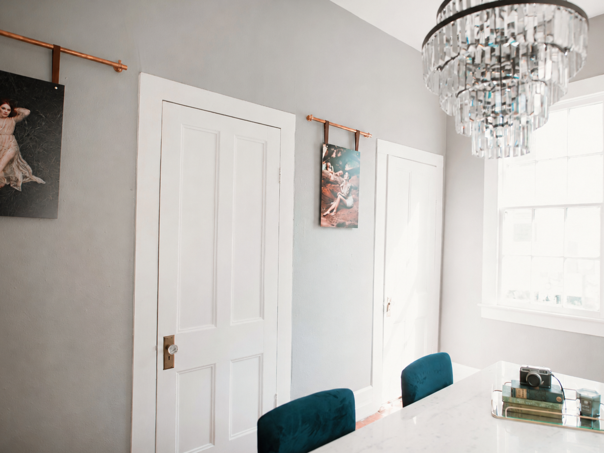 A styled interior room with gray walls, white paneled doors, a crystal chandelier, a dining table, teal chairs, and framed wall art hanging from copper rods.