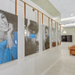 A long hallway with large framed portrait photographs mounted on the wall, including a prominent black-and-white photo of a smiling woman in a blue sweater. The room has light neutral walls, a ceiling-mounted TV, pendant lights, and a brown upholstered chair in the distance.