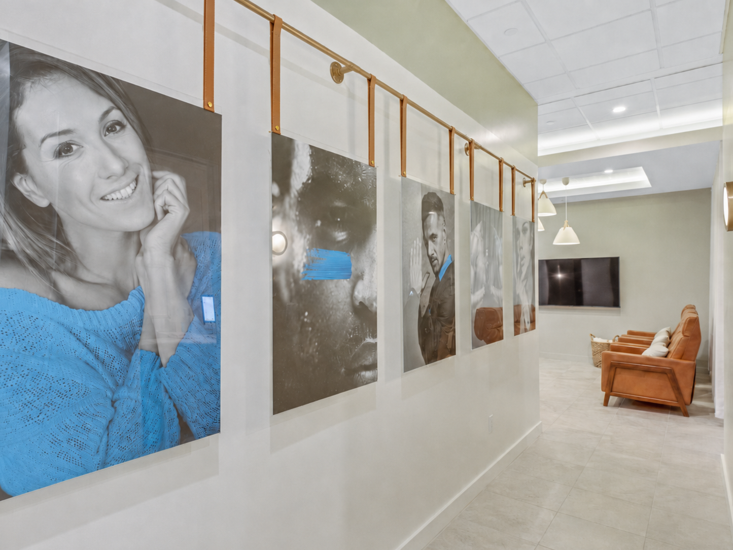 A long hallway with large framed portrait photographs mounted on the wall, including a prominent black-and-white photo of a smiling woman in a blue sweater. The room has light neutral walls, a ceiling-mounted TV, pendant lights, and a brown upholstered chair in the distance.
