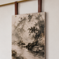 A rectangular canvas wall art print featuring a monochrome landscape painting of trees and rocks by a river, hanging from dark brown leather straps on a brass rod against a light wall.