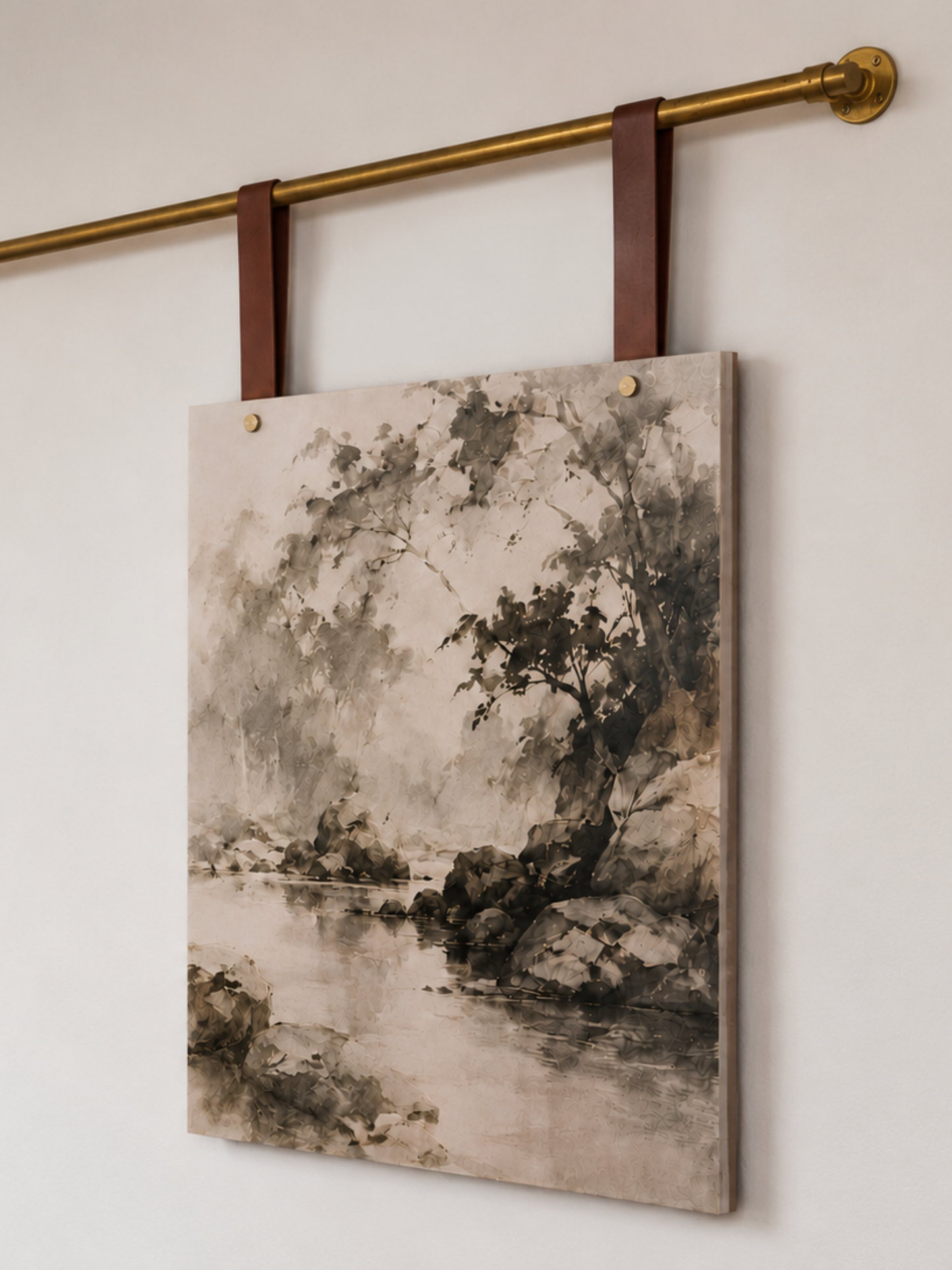 A rectangular canvas wall art print featuring a monochrome landscape painting of trees and rocks by a river, hanging from dark brown leather straps on a brass rod against a light wall.