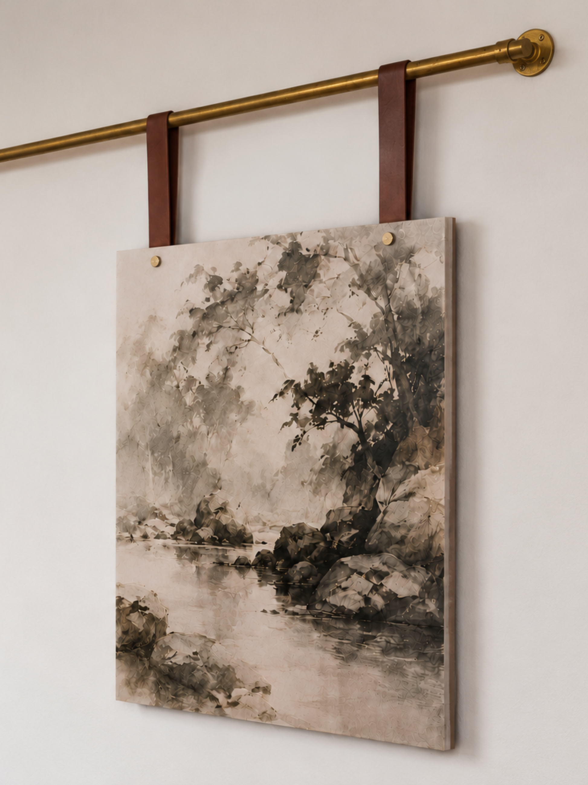 A rectangular canvas wall art print featuring a monochrome landscape painting of trees and rocks by a river, hanging from dark brown leather straps on a brass rod against a light wall.
