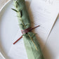 Keyaiira Leather Napkin Tie in mahogany brown wrapped around a green linen napkin with a sprig of greenery, sustainable byproduct leather cord adds a warm, minimalist touch to modern table settings.