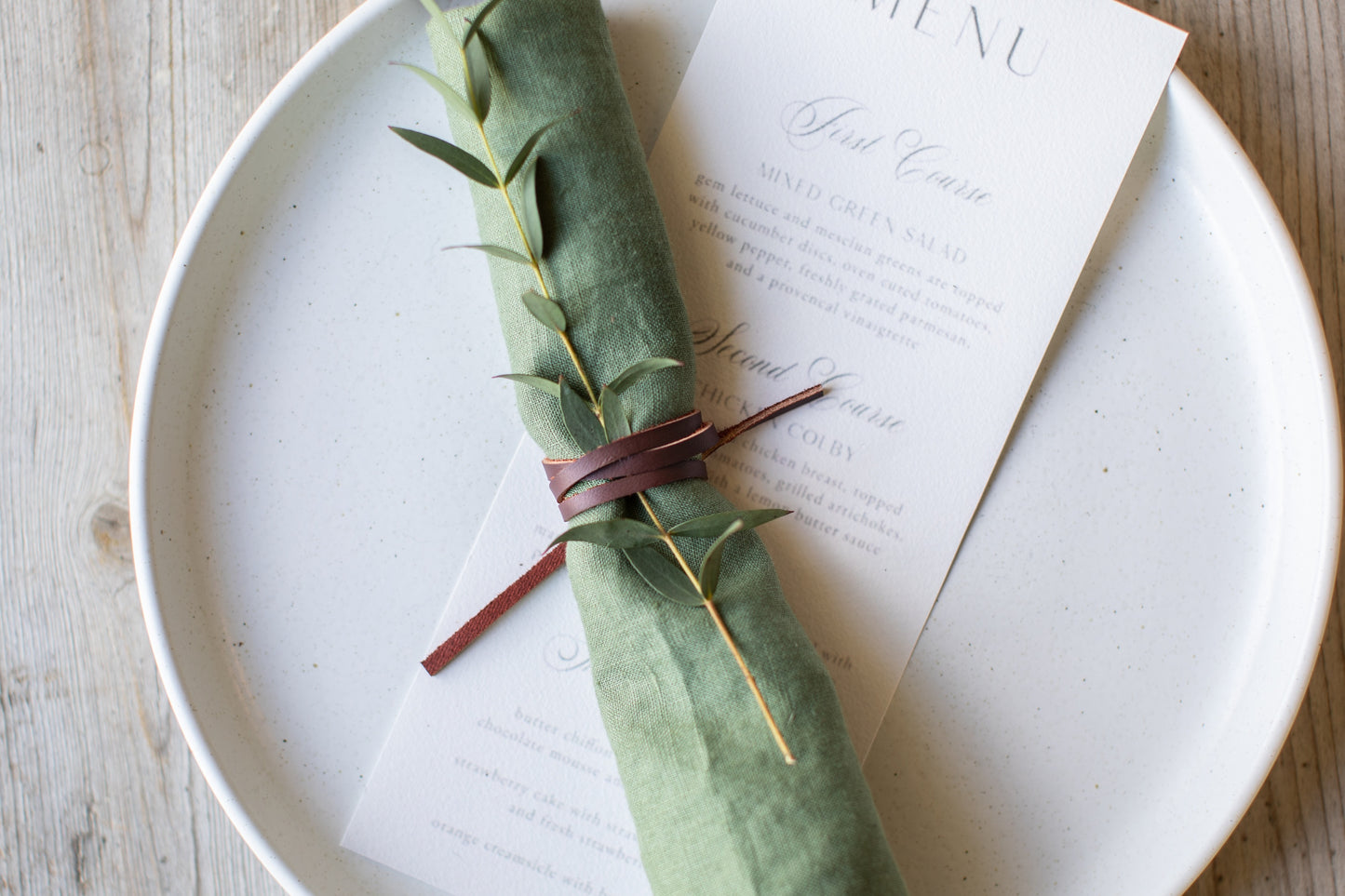 Keyaiira Leather Napkin Tie in mahogany brown wrapped around a green linen napkin with a sprig of greenery, sustainable byproduct leather cord adds a warm, minimalist touch to modern table settings.