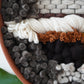 Close view of cream twisted wool layered over dark felted fibers, rust colored yarn and woven leather.