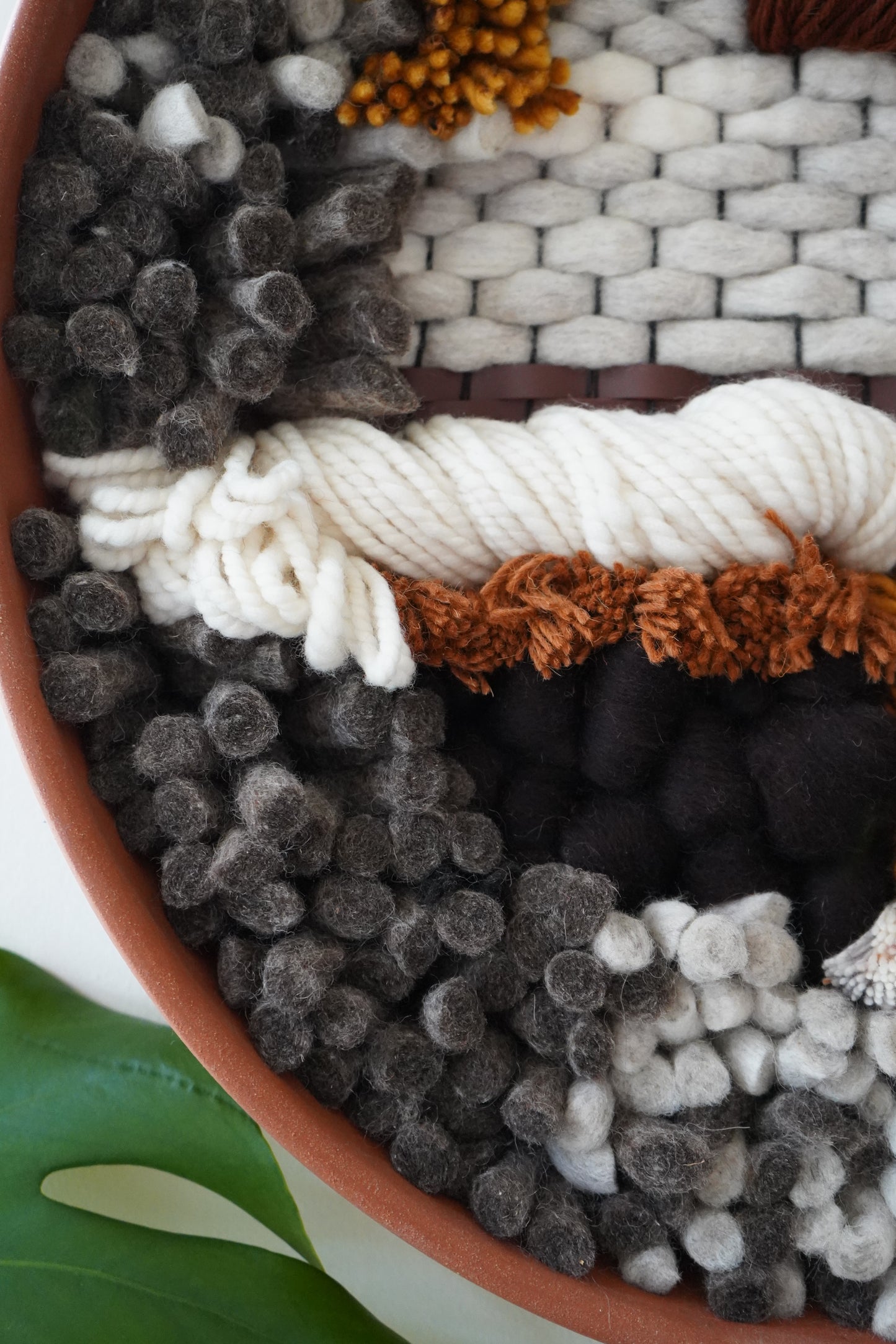 Close view of cream twisted wool layered over dark felted fibers, rust colored yarn and woven leather.