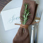 Keyaiira Leather Napkin Tie in Nicotine leather shown with a brown linen napkin, rosemary sprig, and handwritten place card, handcrafted from sustainable byproduct leather for earthy, elegant table settings