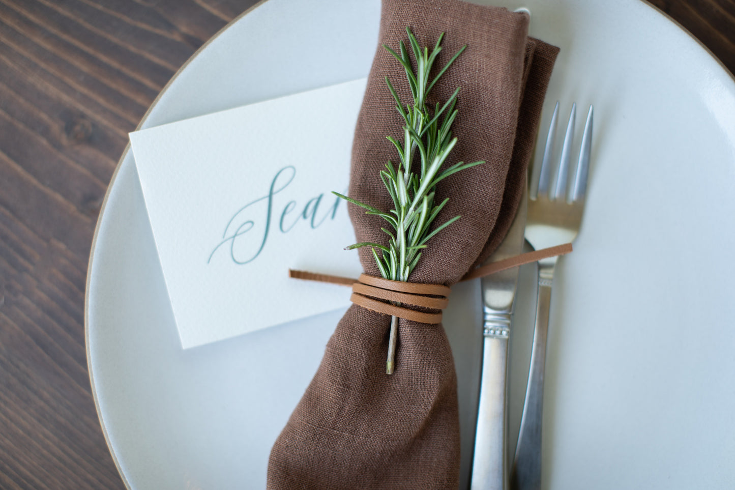Keyaiira Leather Napkin Tie in Nicotine leather shown with a brown linen napkin, rosemary sprig, and handwritten place card, handcrafted from sustainable byproduct leather for earthy, elegant table settings