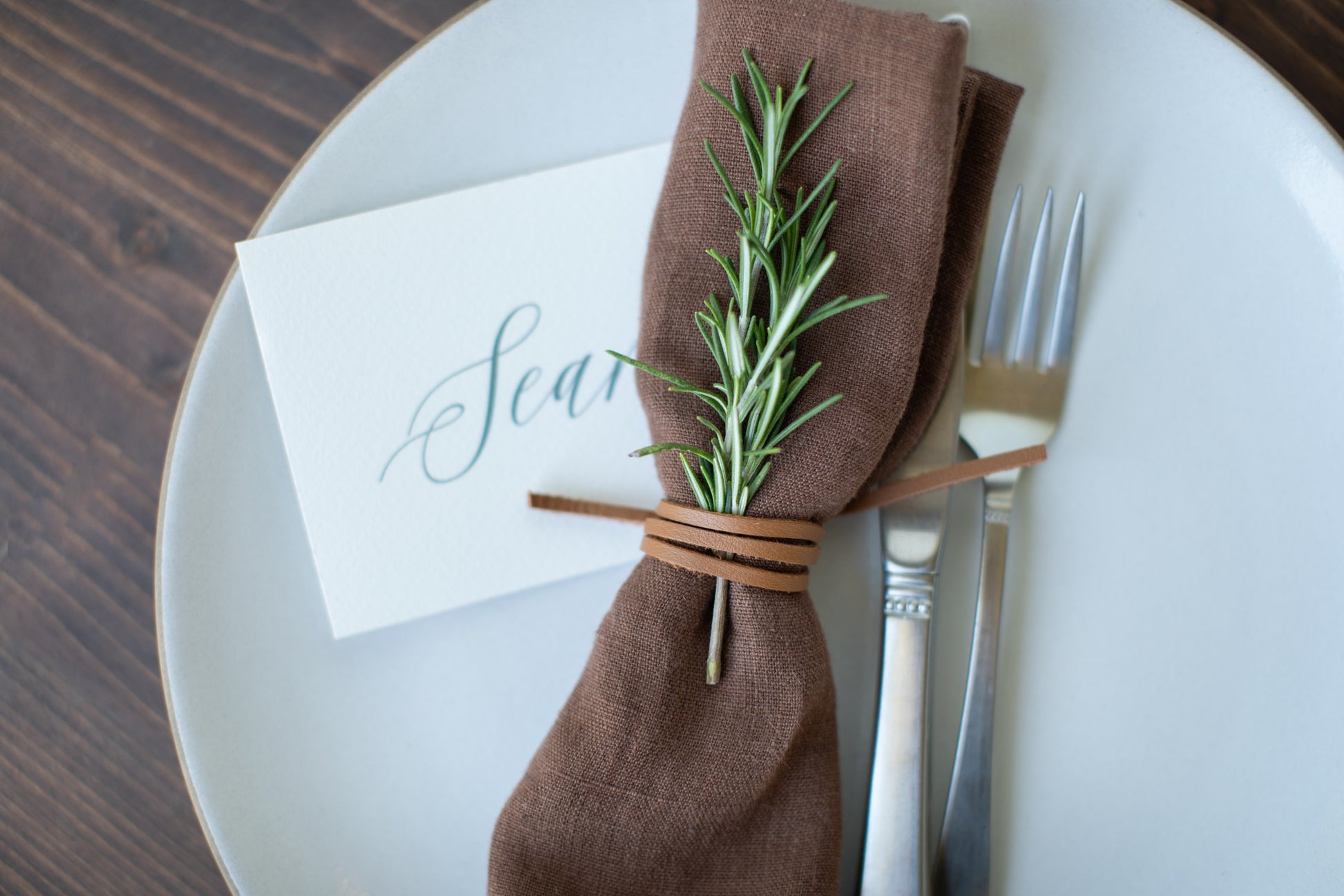 Keyaiira Leather Napkin Tie in Nicotine leather shown with a brown linen napkin, rosemary sprig, and handwritten place card, handcrafted from sustainable byproduct leather for earthy, elegant table settings