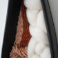 Close angle of the warm brown thick and thin yarn, creamy Rambouillet wool, and hand dyed fibers layered inside the black clay frame. Focused on the natural texture and organic materials.