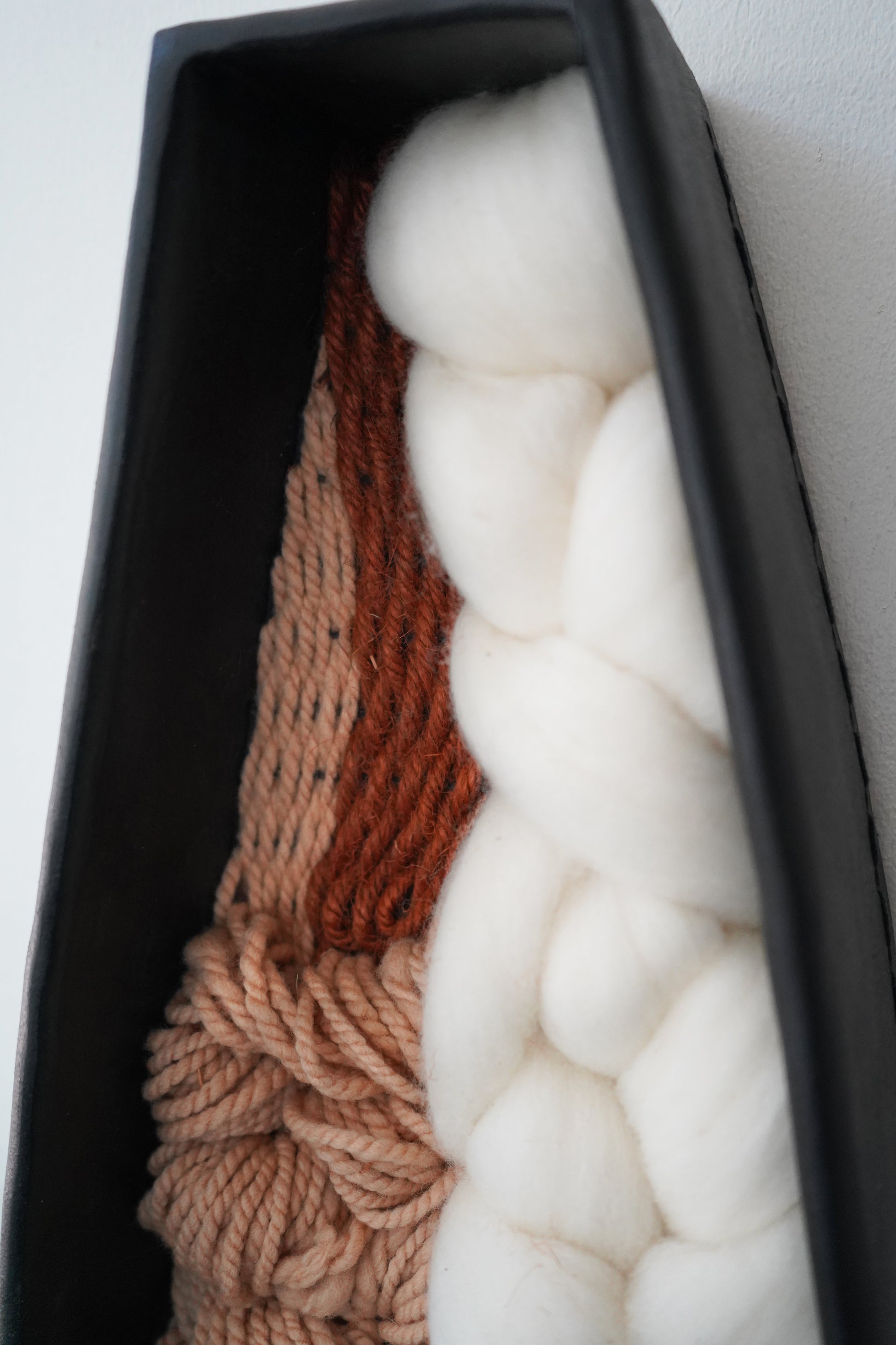 Close angle of the warm brown thick and thin yarn, creamy Rambouillet wool, and hand dyed fibers layered inside the black clay frame. Focused on the natural texture and organic materials.