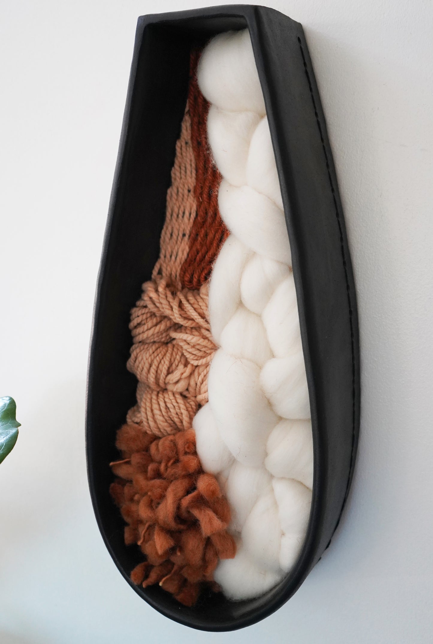 Angled view of the full sculptural piece showing the curvature of the terracotta clay frame and the combination of soft white wool, warm brown yarn, and avocado dyed fibers. Textural fiber and clay art for modern interiors.