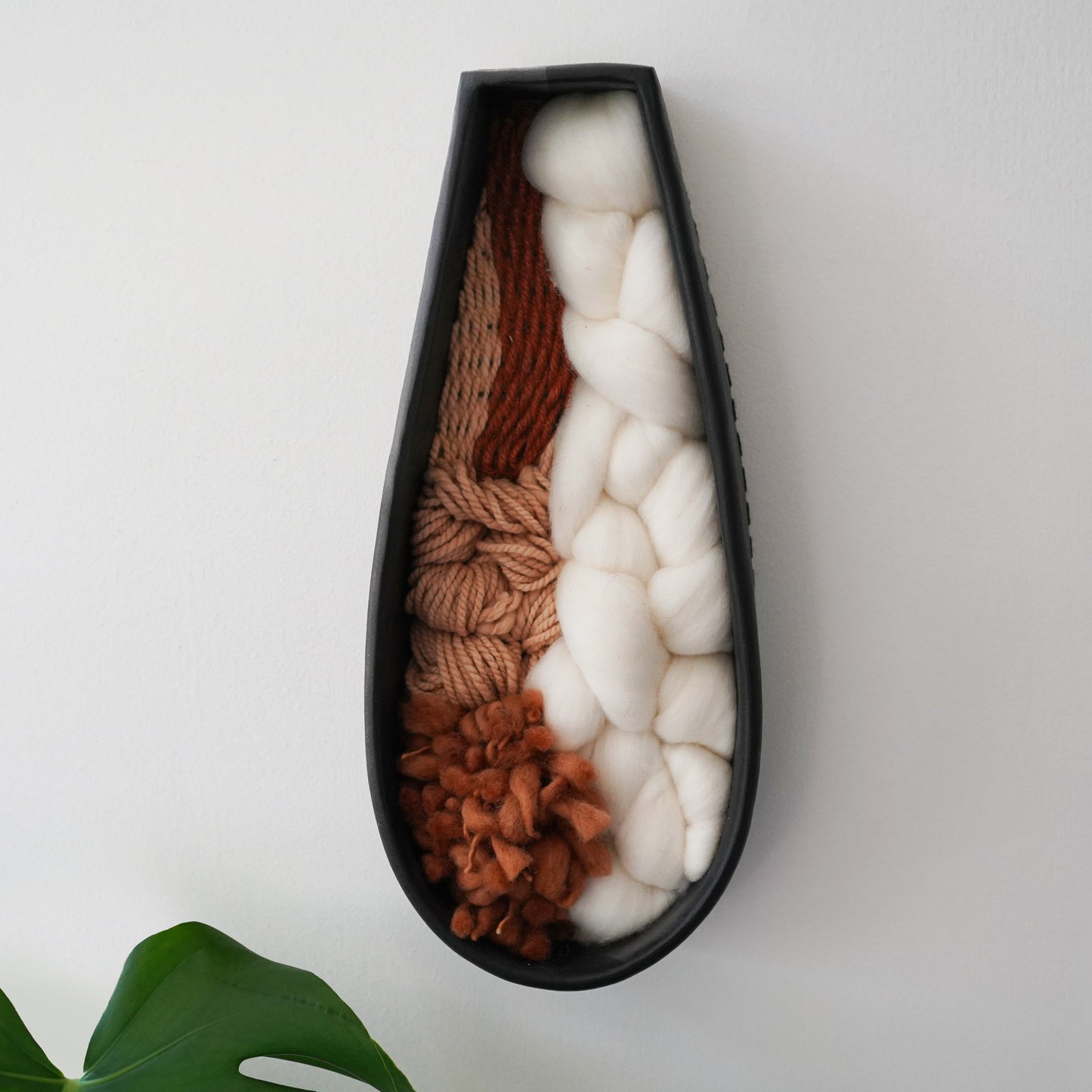Elongated black terracotta clay wall hanging with softly layered Targhee wool, Rambouillet roving, and hand spun thick and thin yarn in warm browns and creamy whites. Modern sculptural fiber and clay artwork photographed against a clean wall with a green plant in the corner.