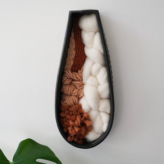 Elongated black terracotta clay wall hanging with softly layered Targhee wool, Rambouillet roving, and hand spun thick and thin yarn in warm browns and creamy whites. Modern sculptural fiber and clay artwork photographed against a clean wall with a green plant in the corner.