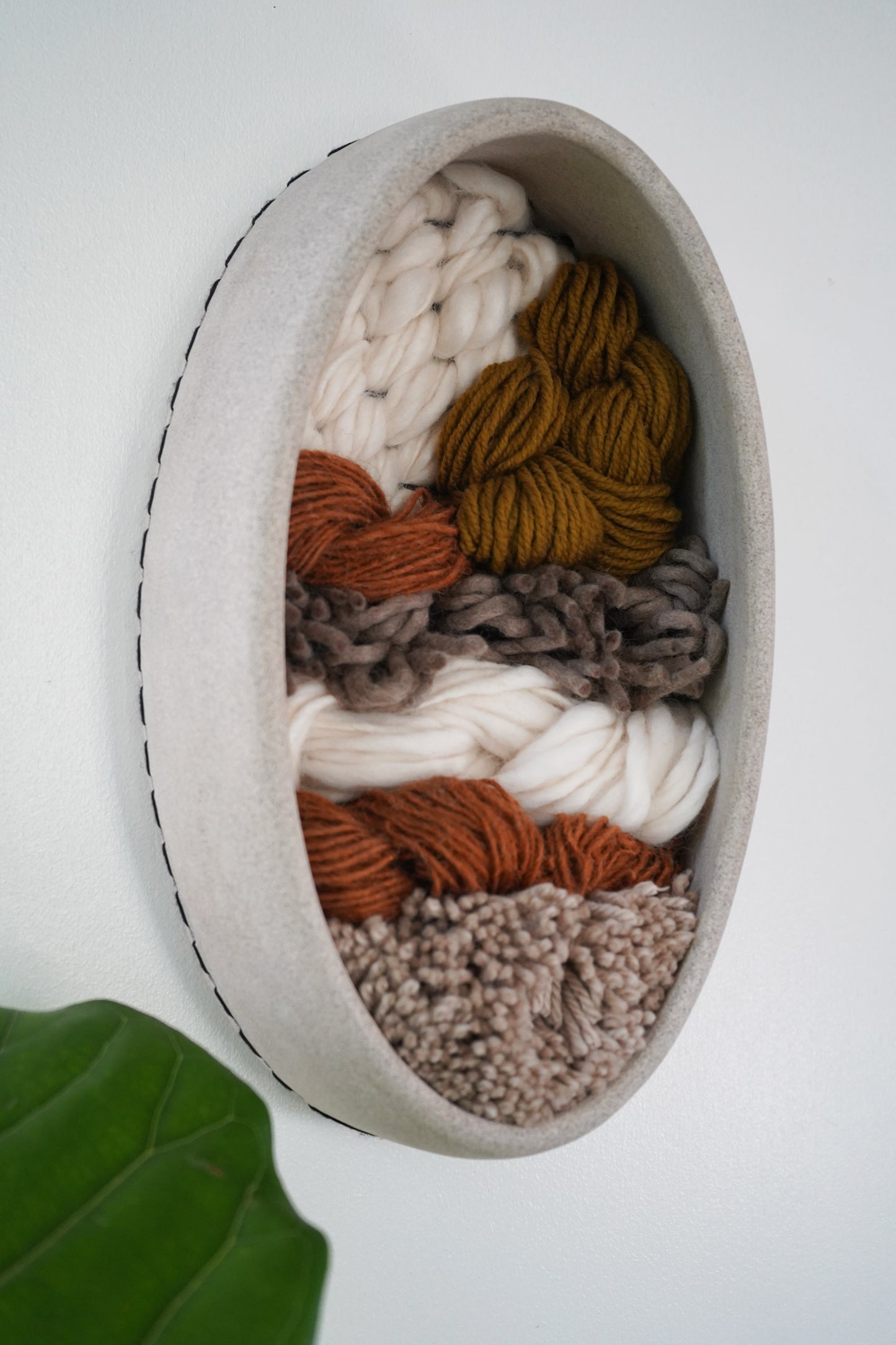 Oval-shaped Handcrafted clay and fiber wall sculpture with woven wool layers in natural tones, contemporary neutral wall art for home or boutique spaces on a white background