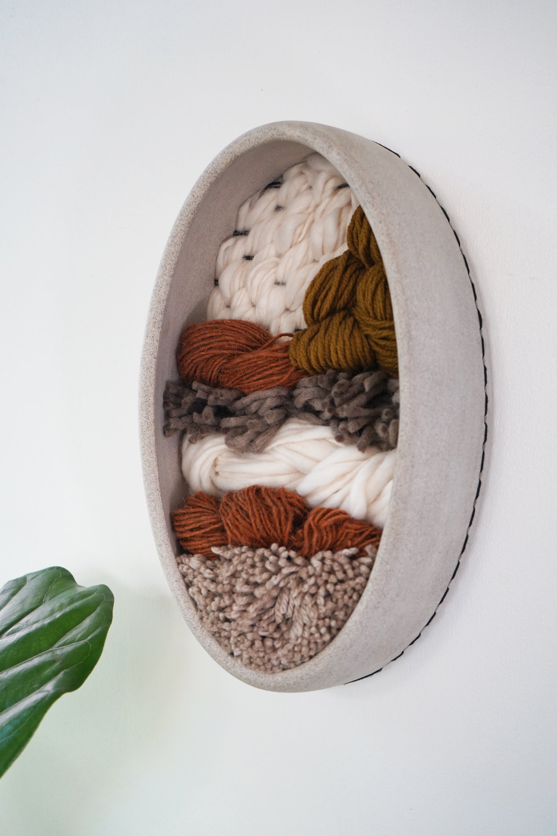 Egg-shaped, sustainable fiber wall sculpture framed in gray clay with warm, earthy textures, handmade modern decor inspired by natural materials