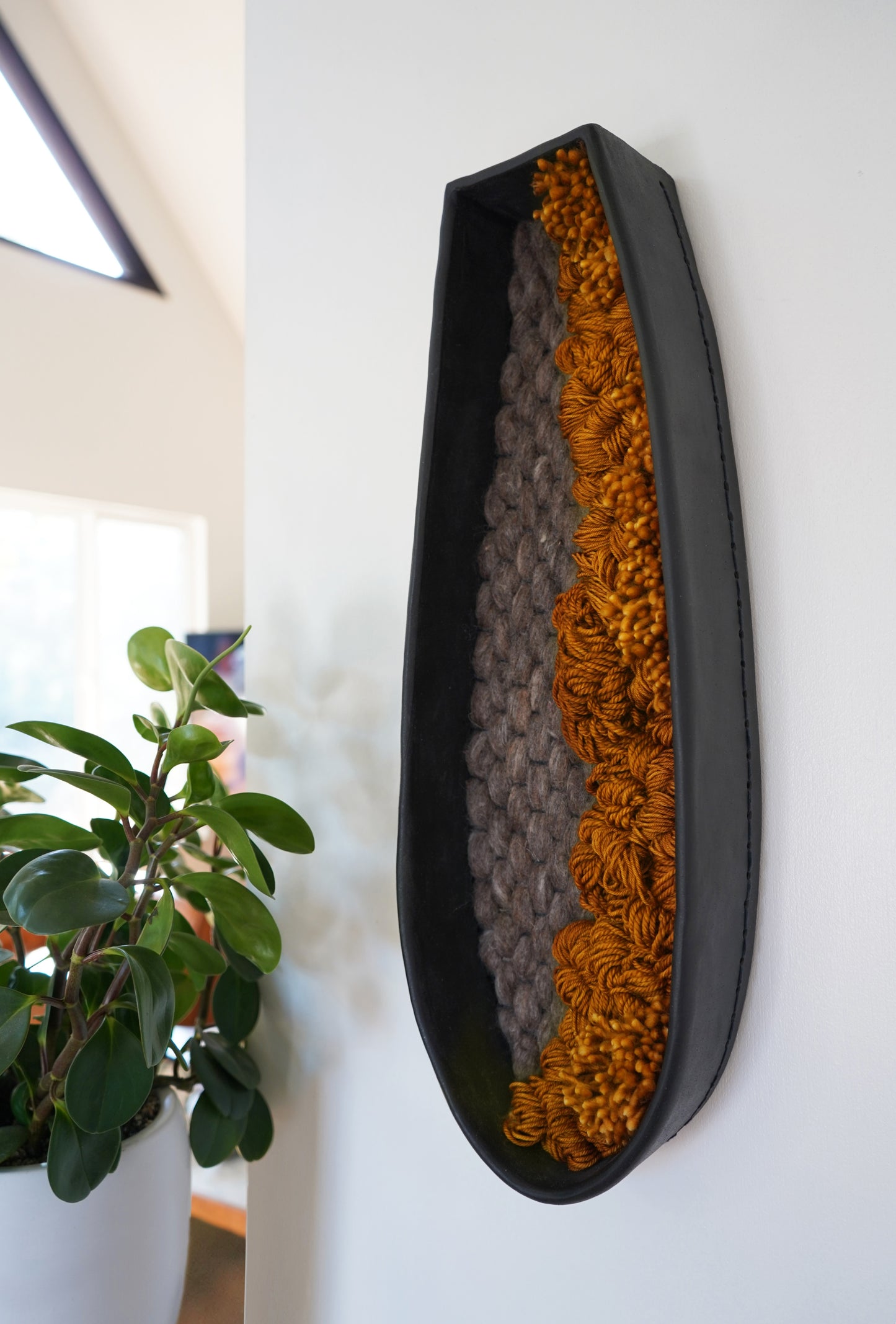 Interior design styling showing textural wool art next to fresh greenery. High-contrast black ceramic and mustard fiber wall hanging for residential or commercial projects.