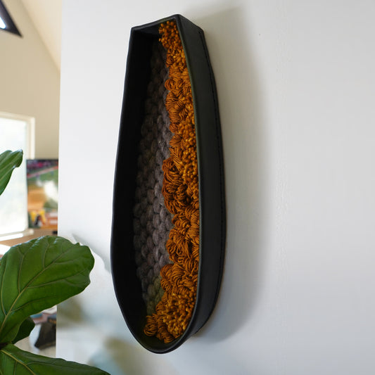 Modern fiber art wall hanging with rusted orange and gray wool framed in long black clay, styled against a white wall with greenery.